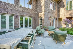 A patio with a wooden table and chairs, a brick building, and a small garden with a purple flowering plant.