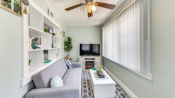 Un salon avec un canapé gris, des étagères blanches, un téléviseur à écran plat, un ventilateur de plafond et un sol à motifs.