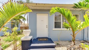 Une maison avec une porte blanche, un petit porche et un jardin planté de palmiers.