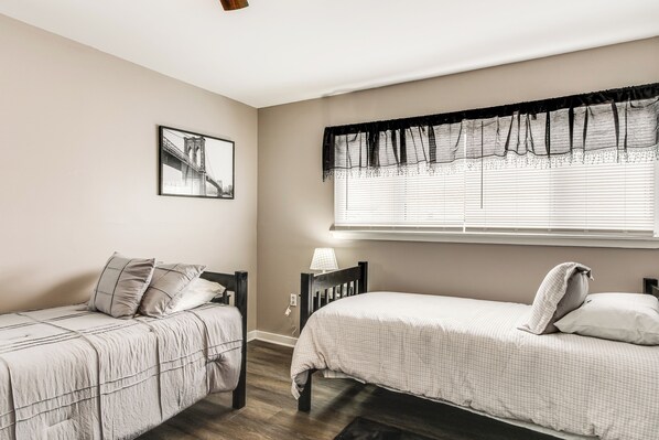 A bedroom with two beds, a window with blackout curtains, a bedside table with a lamp, and a framed picture on the wall.