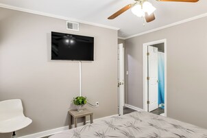 A bedroom with a bed, a TV mounted on the wall, a small wooden side table with a plant, a white chair, and a ceiling fan.