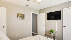 Un dormitorio con cama, ventilador de techo, televisor montado en la pared y una mesita con una planta.