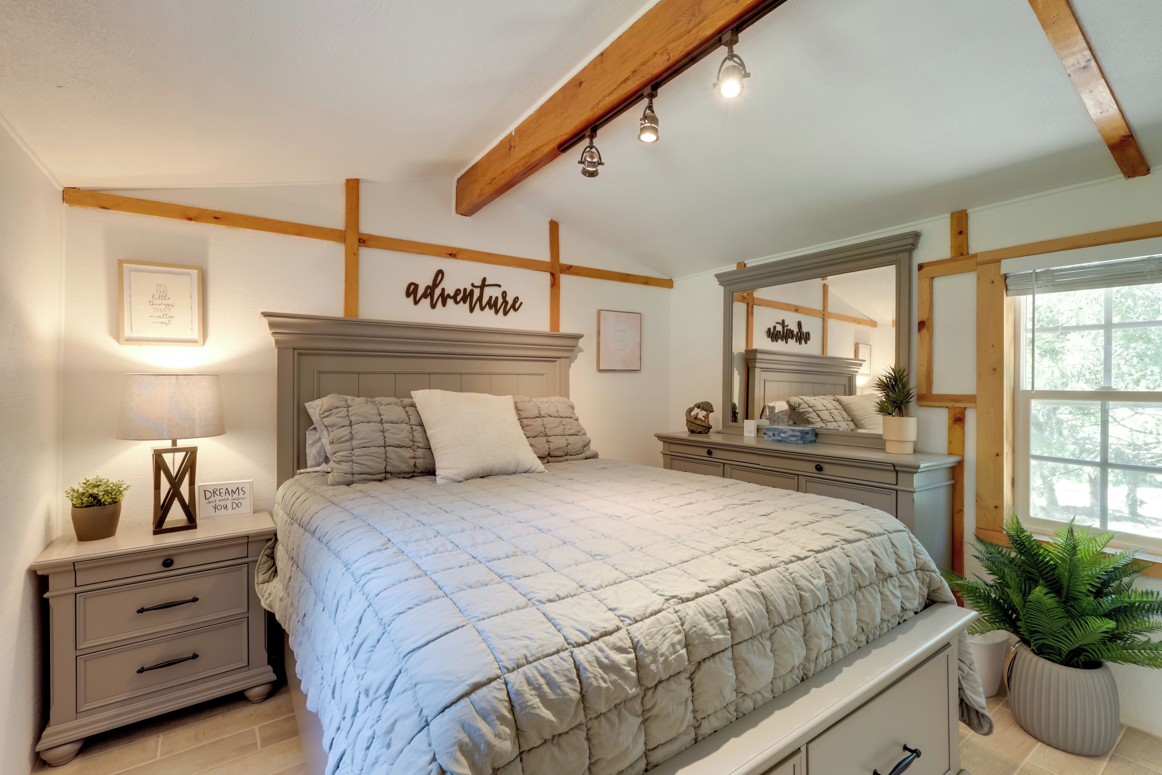 Une chambre à coucher avec un lit, des tables de chevet, un miroir, un mur sur lequel est écrit le mot « aventure », et une fenêtre avec une plante.