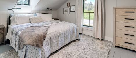 A bedroom with a sloped ceiling, a bed with patterned bedding, a nightstand, a dresser, and a window with curtains.
