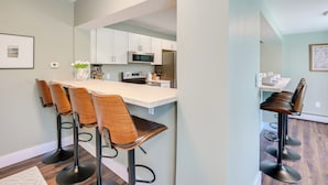 A modern kitchen with white cabinets, a stainless steel refrigerator, and a microwave. There are four wooden bar stools with metal bases. - Pond Views: Spacious Family Home in Presque Isle! (Presque Isle)