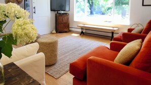 Un salon avec un canapé, un téléviseur, une fenêtre donnant sur des arbres et un vase rempli de fleurs.