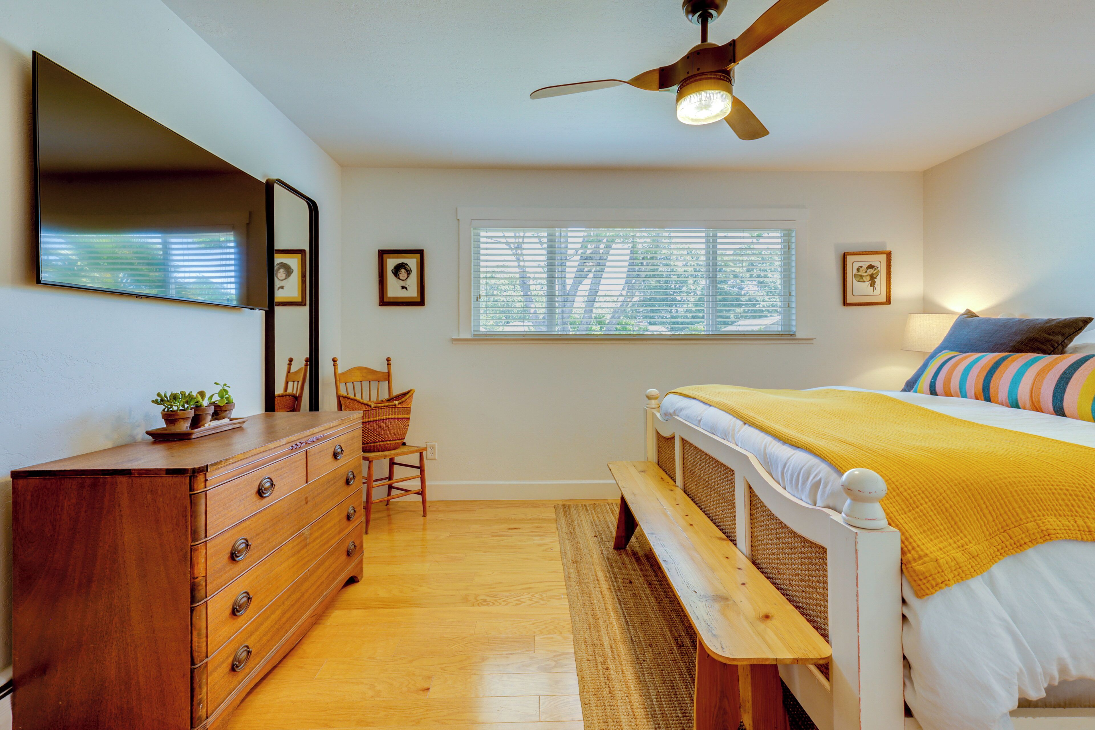 Une chambre avec une commode en bois, un lit recouvert d’une couverture jaune, un ventilateur de plafond et une fenêtre avec des stores.