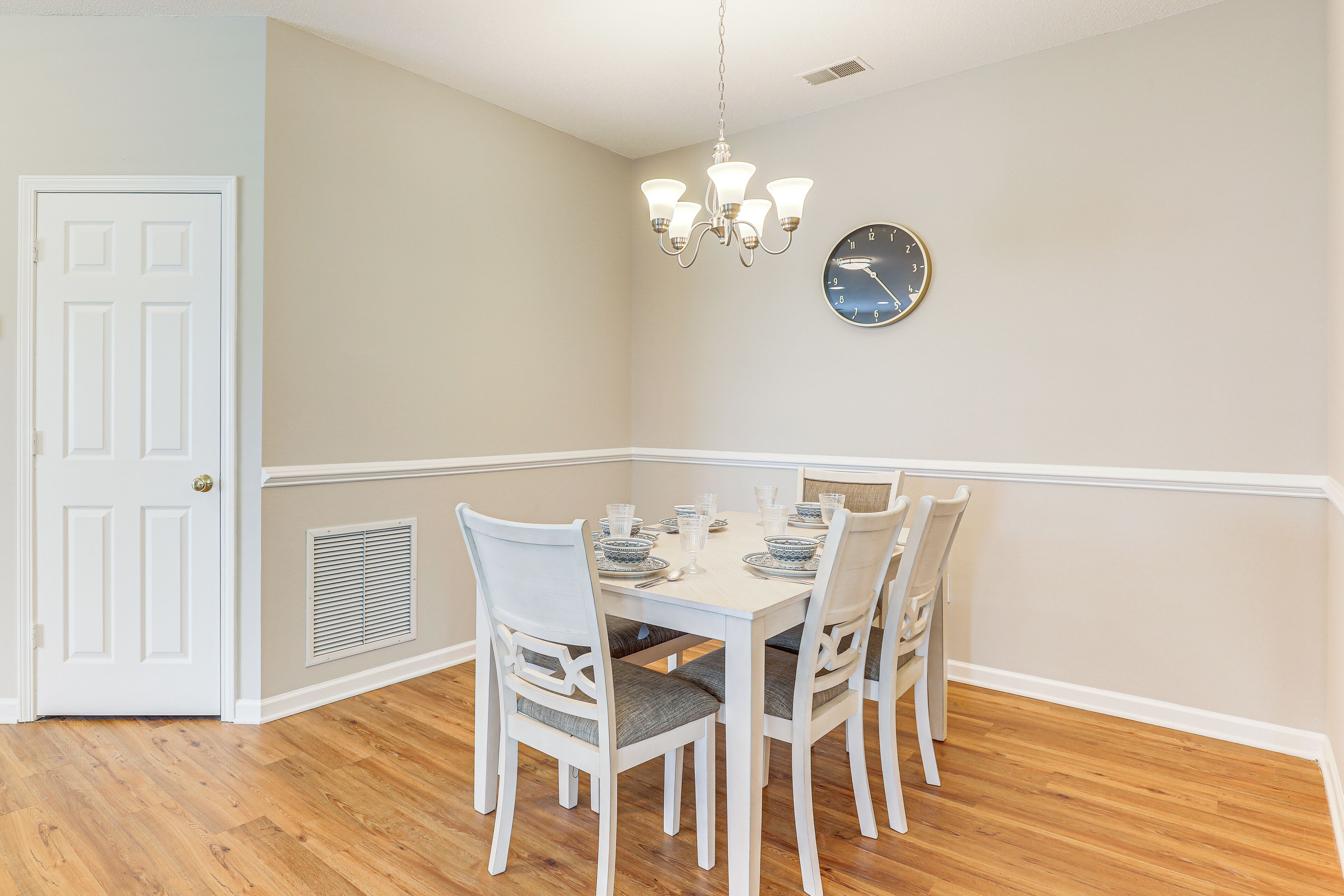 Un coin repas avec une table blanche dressée pour quatre personnes, entourée de chaises blanches dotées de coussins gris. Un lustre est suspendu au plafond et une horloge est fixée au mur.