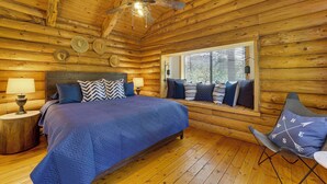 Ein Schlafzimmer in einer Blockhütte mit einem Bett, einem Fenster mit Jalousien, einem Deckenventilator und einem Sessel mit Kissen.