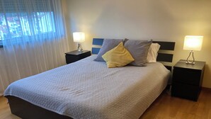 Un lit bien fait, recouvert d’une couverture grise, deux tables de chevet avec des lampes et une fenêtre ornée de rideaux légers.