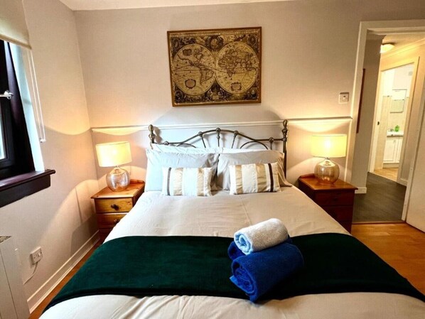 Ein ordentlich gemachtes Bett mit grüner Decke, weißen Kissen und einem zusammengerolltem Handtuch. Zwei Nachttischlampen und ein an der Wand befestigtes Globus-Kunstwerk sind vorhanden.