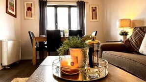 Un salon chaleureux avec une table basse en bois, une bougie allumée, un chandelier en verre et une plante en pot. On y trouve également une table à manger avec des chaises et une fenêtre avec des rideaux.