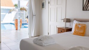 A hotel room with a bed, a nightstand, a lamp, a door, and a view of an outdoor swimming pool. - Ocean Whisper Villa in Sint Willibrordus (St. Marie)
