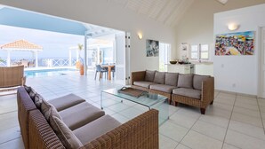 A spacious living area with a glass coffee table, wicker sofas, and a view of a pool and outdoor seating. - Ocean Whisper Villa in Sint Willibrordus (St. Marie)
