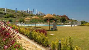 A pool area with a grassy lawn, thatched umbrellas, and a clear view of buildings in the background. - Chic Coastal Living on the New Golden Mile (Estepona)