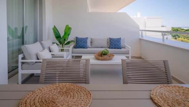 A balcony with a white sofa, two woven poufs, a small table with a plant, and a large window overlooking a cityscape. - Chic Coastal Living on the New Golden Mile (Estepona)