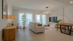 A modern living room with a sofa, a dining table with chairs, a TV mounted on the wall, and a large window with sheer curtains. - Chic Coastal Living on the New Golden Mile (Estepona)