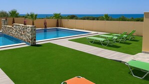 Une piscine bordée d’un mur en pierre et entourée de palmiers, avec de l’herbe artificielle verte et des chaises longues.