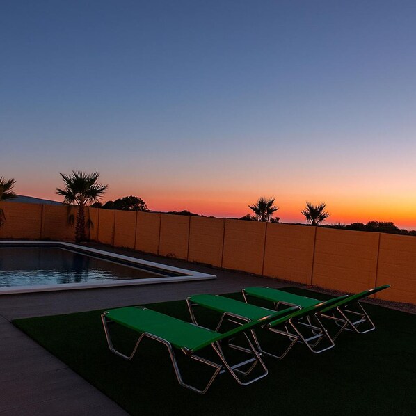 Zona de piscina con vista al atardecer, sillas reclinables verdes y palmeras.