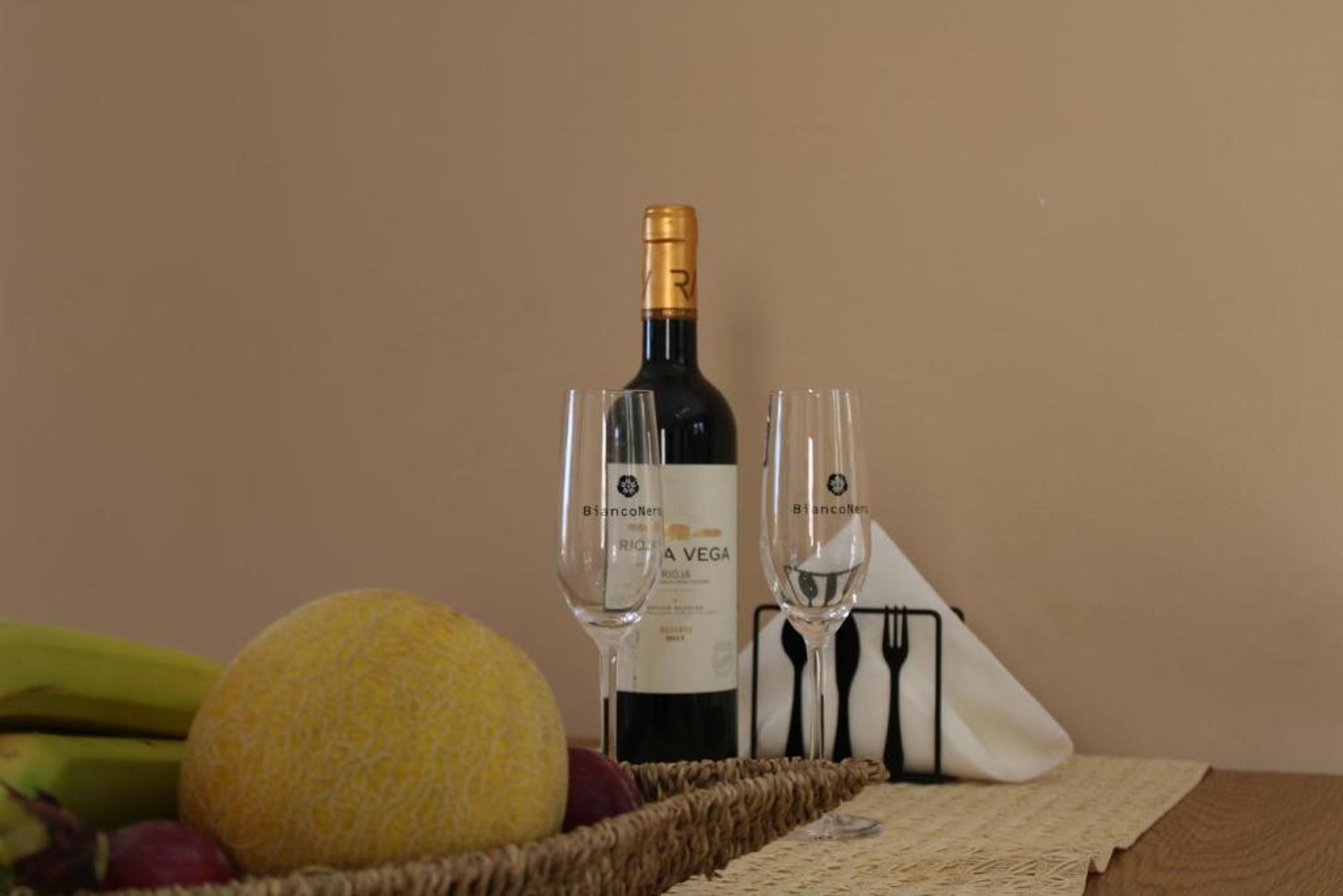 Una ampolla de vi, dues copes, una cistella de fruites i un jeu de draps sobre una taula.
