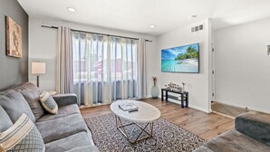 A modern living room with a grey sofa, a round coffee table, and a patterned rug. There is a large window with sheer curtains, a painting on the wall, and a small table with decorative items. - Spacious Family Friendly House in Central Location (La Puente)
