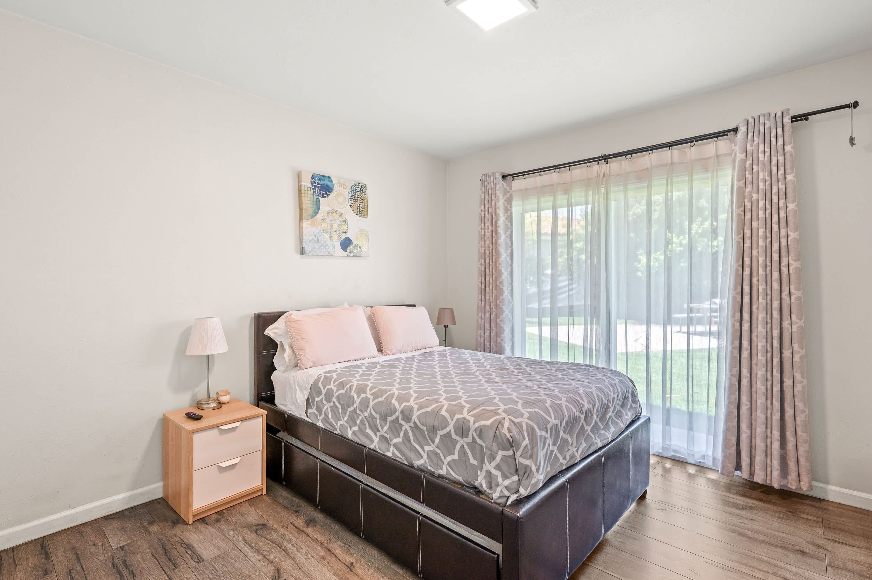 A bedroom with a bed, a nightstand, a lamp, a painting, and a large window with curtains.