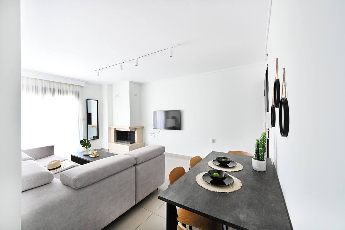 A modern living room with a grey sofa, a dining table with placemats, a flat-screen TV, and a cactus plant.