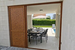 Une porte coulissante ouverte donne accès à un espace repas extérieur agrémenté d’une piscine et d’une haie.