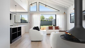 A modern living room with a fireplace, a sofa, a dining area, and large windows. - 13A Alice Street, Jindabyne (Jindabyne)