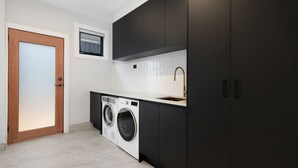 A compact kitchen with a built-in washer and dryer, a small window, and dark cabinetry. - 13A Alice Street, Jindabyne (Jindabyne)