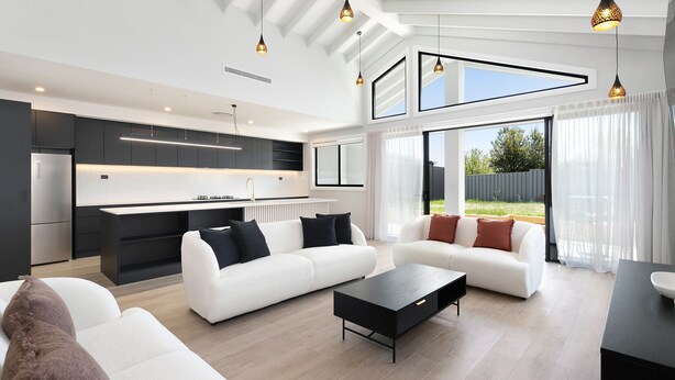 A modern living room with a white sofa, a black coffee table, and a kitchen area with a built-in oven and a large window. - 13A Alice Street, Jindabyne (Jindabyne)