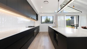 A modern kitchen with dark cabinetry, a white countertop, and a large window providing natural light. - 13A Alice Street, Jindabyne (Jindabyne)