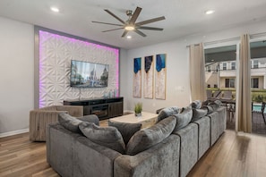 A modern living room with a sectional sofa, a flat-screen TV, decorative wall panels, and a ceiling fan.