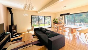 A modern living room with a fireplace, a sofa, a dining area, and large windows offering a view of the outdoors.