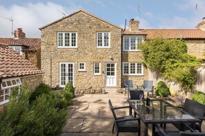 A stone house with a tiled roof, a front door, and a patio with a glass table and chairs. - Robins Cottage - Quaint North Yorkshire cottage. (YORK)