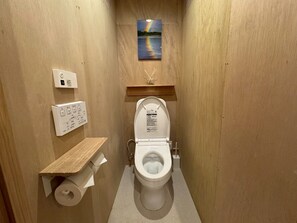 Ein kleines Badezimmer mit Holzoptik, das eine Toilette, eine an der Wand montierte Schalterleiste, einen Toilettenpapierhalter und eine kleine Ablagefläche mit einem Bild umfasst.