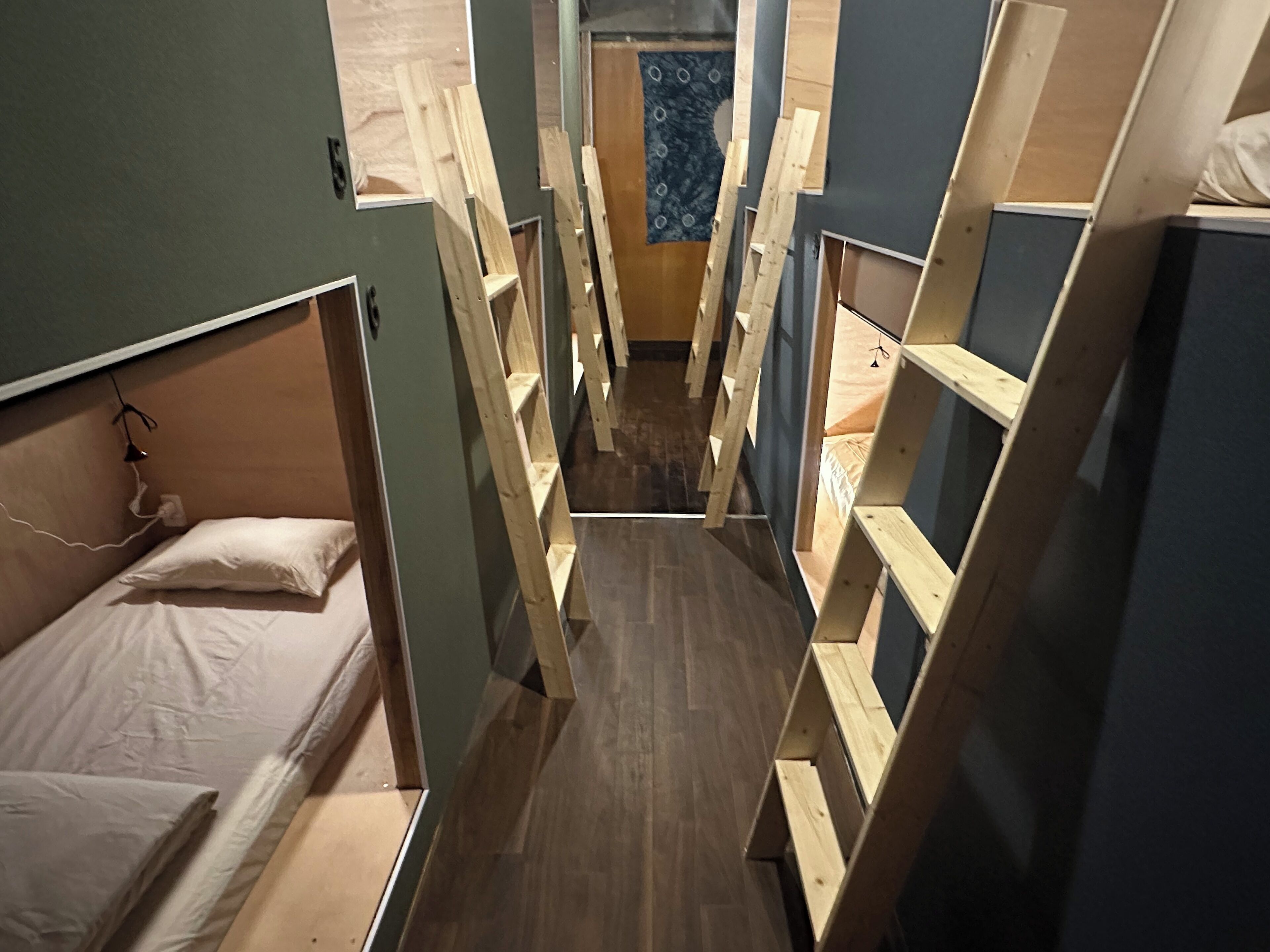 A narrow corridor with bunk beds on either side, wooden flooring, and a blue patterned wall at the end.