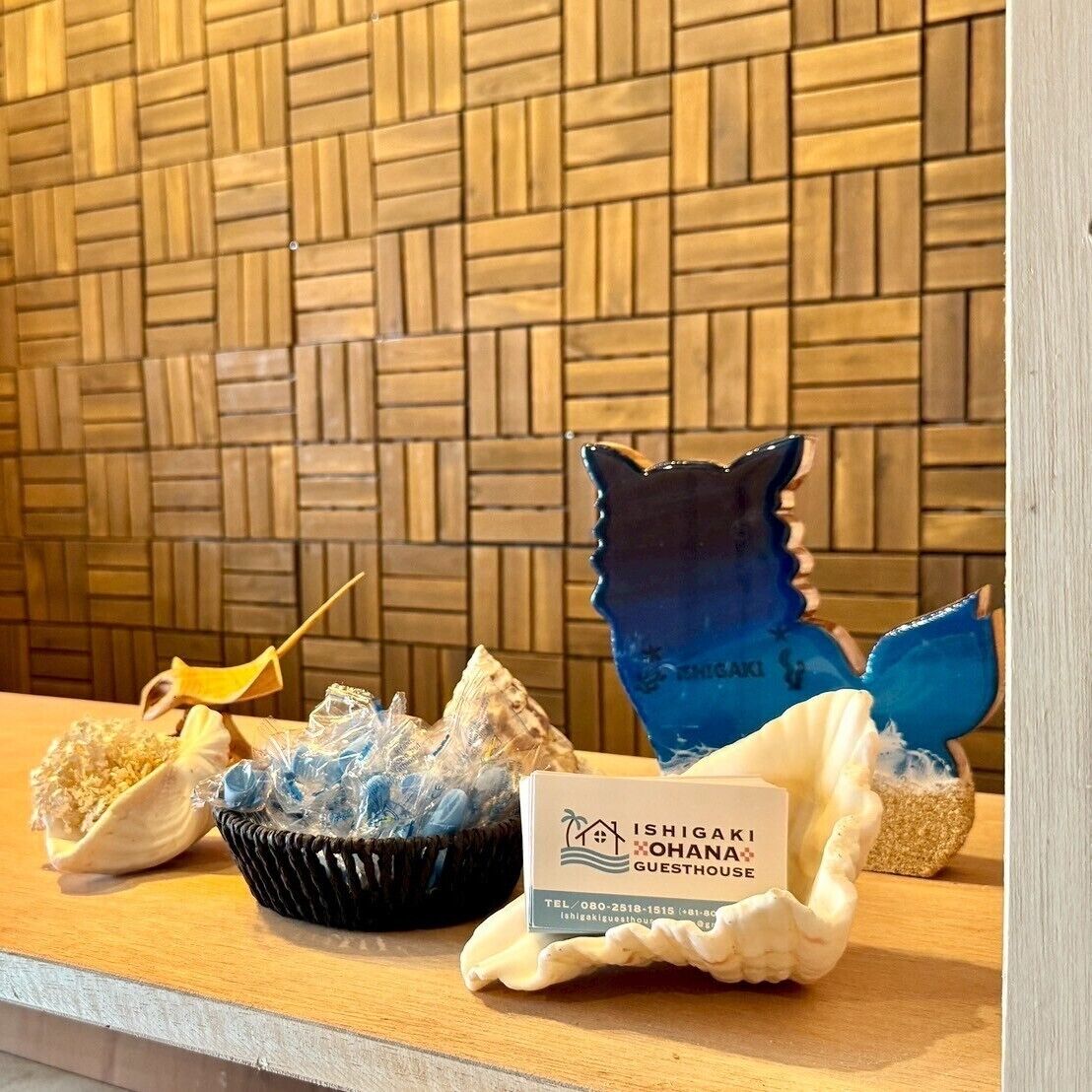 A reception desk with a woven wall background, a blue decorative fish sculpture, a yellow bird figurine, and a sign for Ishigaki Ohana Guesthouse.