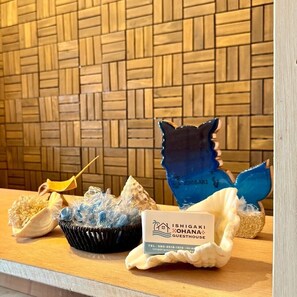 Ein Empfangsschalter mit einer gewobenen Wand, einer blauen Fischskulptur als Dekoration, einer gelben Vogelfigur und einem Schild für die Ishigaki Ohana Guesthouse.
