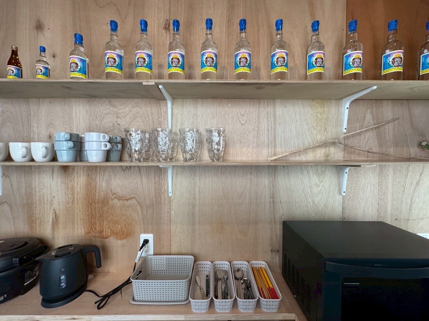 Een keuken met houten planken waarop flessen, bekers en glazen staan, en een aanrecht met een waterkoker, manden en keukengerei.