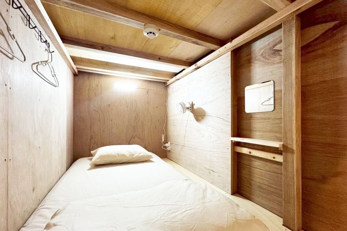 Una habitación pequeña y estrecha con techo y paredes de madera, una cama individual con ropa blanca, una lámpara fijada a la pared y un estante con un objeto blanco.