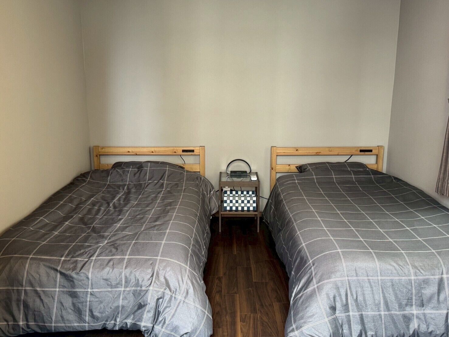 Une chambre avec deux lits simples, chacun doté d’une tête de lit en bois et d’une couette à carreaux gris. Une petite table de chevet avec une radio et un livre est placée entre les lits.