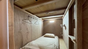 Een kleine kamer met een enkel bed, houten wanden en een schuin dak. Er staat een bed met wit beddengoed en een enkele hanglamp.