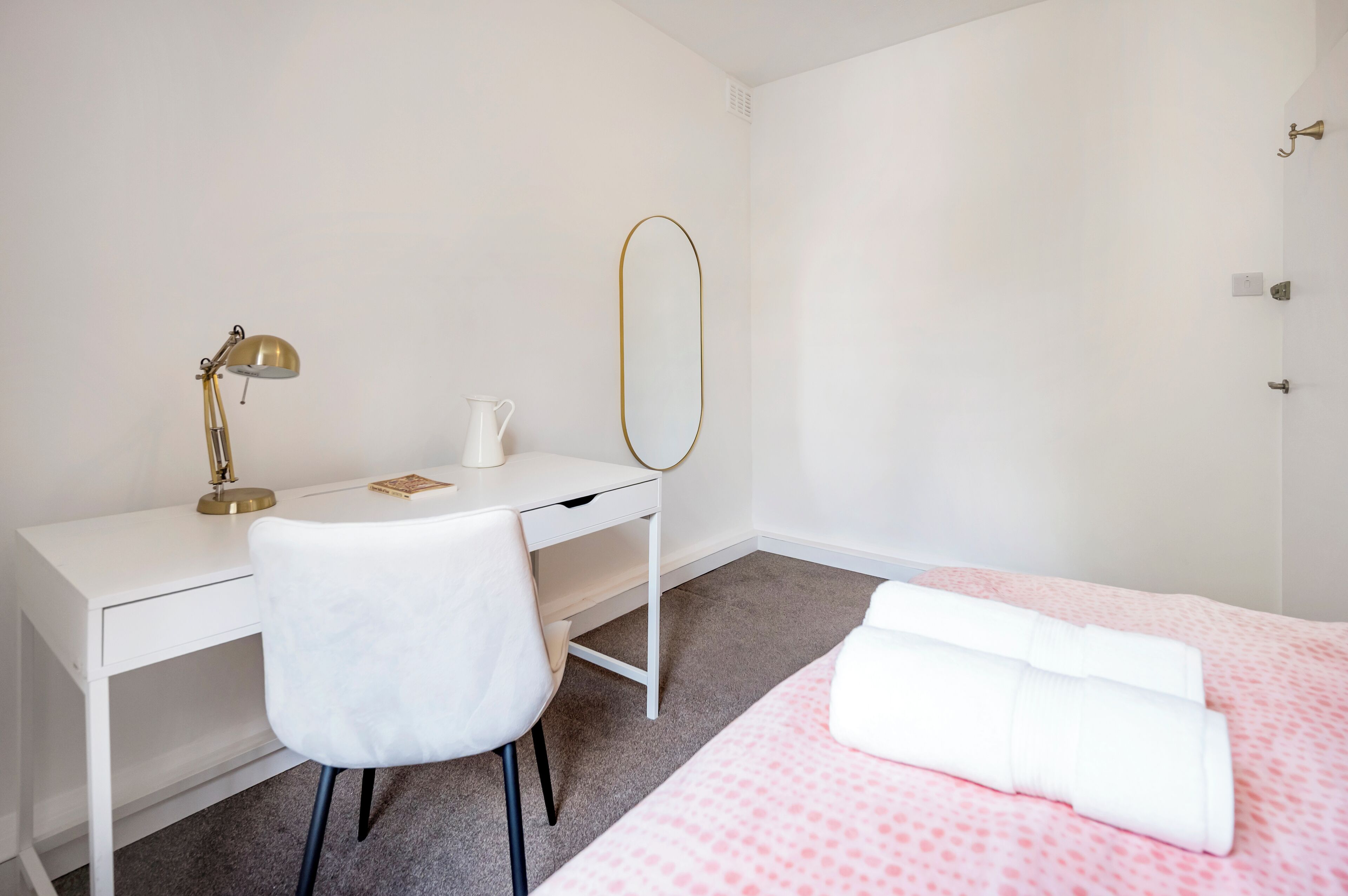 Une chambre avec un bureau blanc, une chaise, un lit avec une literie rose et un miroir avec un cadre doré.