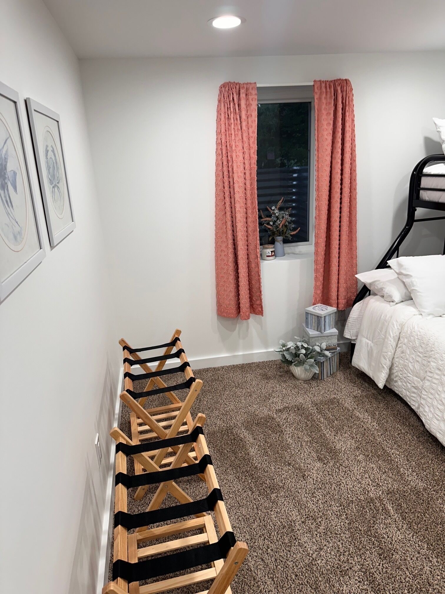 Un dormitorio con una cama litera, un perchero de madera, una ventana con cortinas rosadas y una pequeña planta en el alféizar.