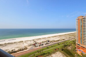 A high-rise hotel overlooking a beach with a clear view of the ocean, a road with parked vehicles, and a parking lot.