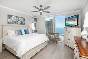 A bedroom with a bed, a television, a ceiling fan, a window with a view of the sea, and a balcony with a wicker chair.