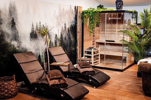 A spa room with two reclining chairs, a wooden massage table, and a glass wall with greenery.