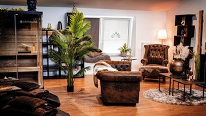A cozy living room with a wooden floor, a brown leather sofa, a tufted armchair, and a large potted plant. - Wellness for two (Ahlen)