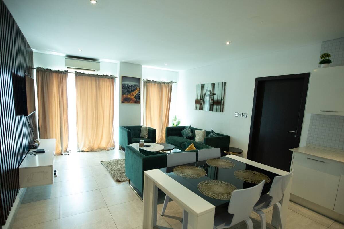 A modern living room with a green sofa, a dining table with chairs, and a kitchen area with white cabinets.